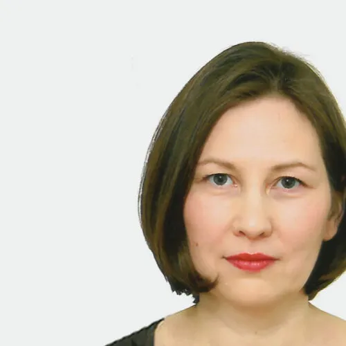 Frau mit kurzen braunen Haaren und rotem Lippenstift vor einem schlichten weißen Hintergrund, in die Kamera blickend.