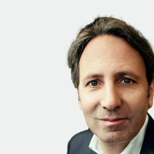 Ein Mann mit dunklem Haar, Anzugjacke und weißem Hemd, der in die Kamera schaut und lächelt.