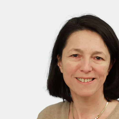 Frau mit schulterlangem dunklem Haar, lächelnd, mit beigem Oberteil, vor einem schlichten hellen Hintergrund.