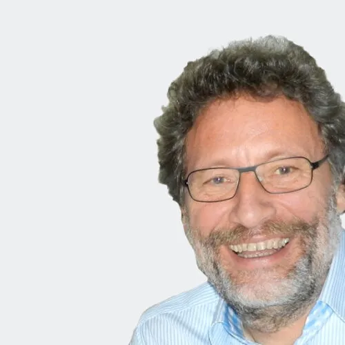 Lächelnder Mann mit Brille und Bart in einem hellblau gestreiften Hemd, vor einem schlichten Hintergrund.